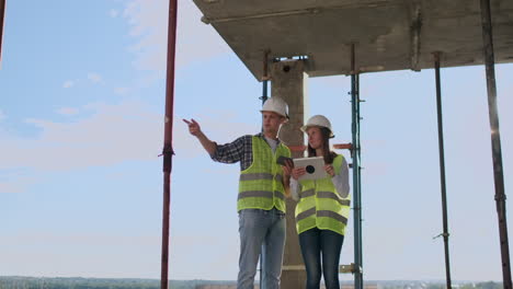 Engineers-builders-man-and-woman-standing-on-the-roof-of-the-building-with-a-tablet-computer-discussing-in-white-helmets-and-shirts.